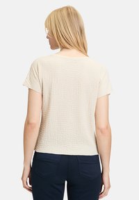 Krótki rękaw, teksturowany beżowy top o luźnym fasonie i okrągłym dekolcie, w połączeniu z granatowymi spodniami, pokazujący widok z tyłu oraz subtelny wzór materiału.