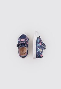 Zapatos de mezclilla azul con patrones florales, acentos rosas y suela de goma blanca. Cierre con tira de velcro. Diseño de punta redondeada y plana.