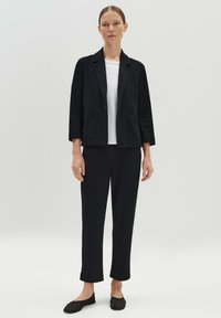 Zwarte, op maat gemaakte blazer met een kraag, twee voorzakken en driekwartmouwen, gedragen over een wit shirt en zwarte broek, met zwarte flats.