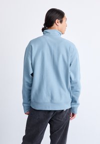Hombre con el pelo recogido, vistiendo un sudadera de cuello alto de color azul claro y unos pantalones vaqueros gris oscuro, de espaldas frente a un fondo blanco liso.