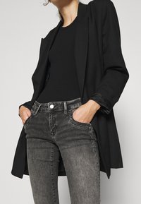 Musta bleiseri mustan topin päällä, yhdistettynä harmaisiin farkkuihin, joissa on hienovaraiset haalistumat, taskut ja metalliset nappikoristeet.