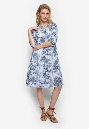 Frau mit blondem Haar in einem knielangen blau-weißen Kleid mit botanischen und tierischen Motiven, die braune flache Riemchensandalen trägt und lässig steht.
