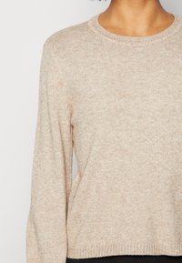 Pull beige en maille avec un col rond, des manches longues et une finition texturée. Le tissu semble doux et légèrement chiné.
