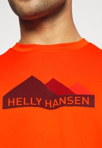 Jasně oranžové tričko s potiskem loga s horami a nápisem "HELly HANSEN" tmavě hnědou barvou. Tkanina vypadá hladká a lehká.