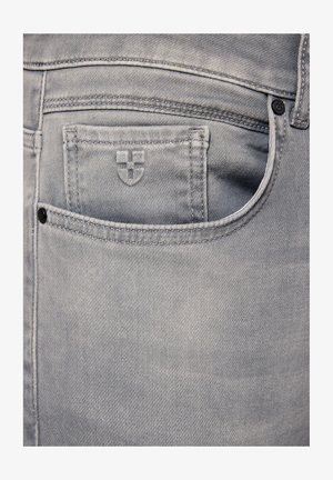 Zbliżenie na jasnoszare dżinsy, ukazujące tylną kieszeń z małym emblematem tarczy i starannym przeszyciem.