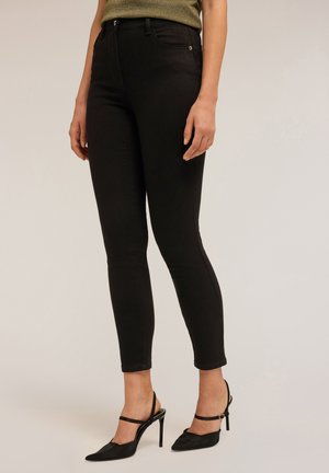 Femme portant un jean skinny noir et des escarpins à talons hauts noirs, se tenant devant un fond neutre et uni.