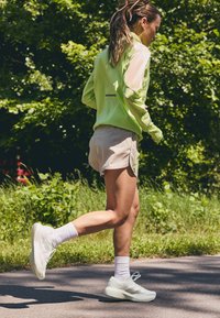 Giacca verde chiaro, con texture e dettagli riflettenti, pantaloni beige e scarpe da ginnastica bianche. Il corridore si trova su un sentiero circondato dal verde.