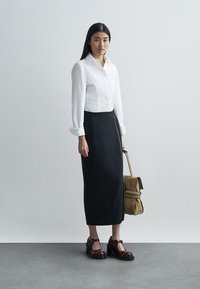 Chemise blanche à boutons, manches longues, jupe crayon noire jusqu'aux genoux, chaussures à plateforme bordeaux et sac en bandoulière marron.