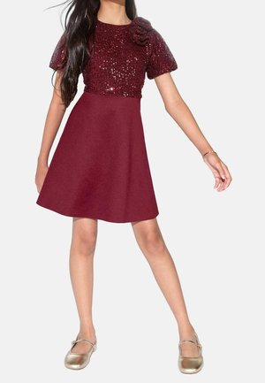 Niña con vestido burdeos con parte superior de lentejuelas y mangas abullonadas, combinado con zapatos Mary Jane dorados, de pie con los brazos y las piernas separados.