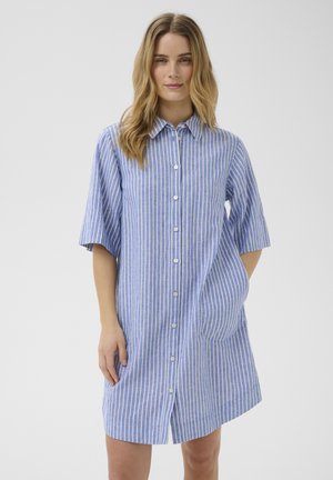 Femme portant une robe chemise à rayures verticales bleu et blanc avec manches courtes, debout devant un fond uni gris clair.