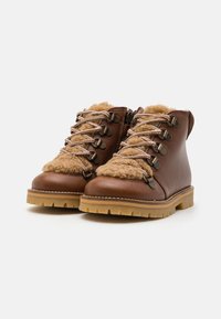 Botas de cuero marrón hasta el tobillo con forro de pelaje sintético, ojales de metal y cordones en contraste. Suela de goma gruesa con patrones de relieve para mayor tracción.