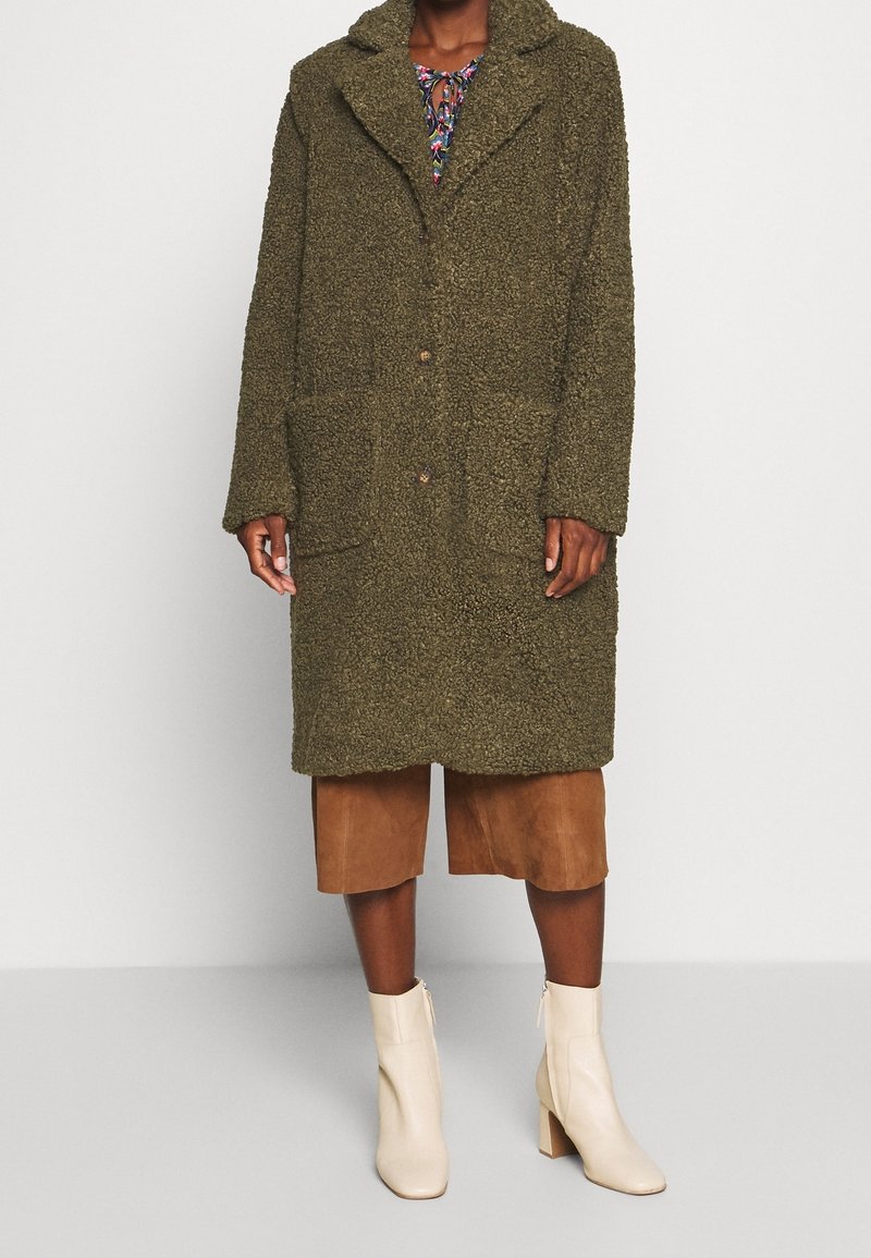 Olivengrøn, struktureret, overdimensioneret frakke med hætte, to frontlommer og knaplukning, båret over brune shorts og beige ankelstøvler.