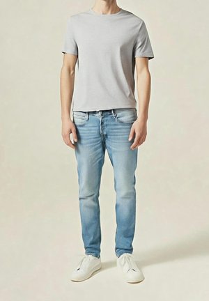 Man staat en draagt een lichtgrijs T-shirt met korte mouwen, lichtblauwe spijkerbroek en witte sneakers tegen een neutrale achtergrond.