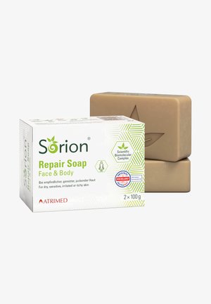 Reparatur-Seife in einer verpackten Box mit grünen Akzenten, zwei beigen rechteckigen Seifenstücken mit einem geprägten Blätterdesign. Geeignet für empfindliche Haut.