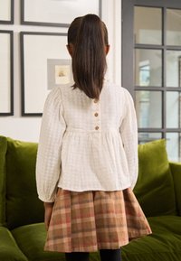 White textured blouse with puffed sleeves and back buttons, paired with a plaid brown and red skirt, worn over black tights.