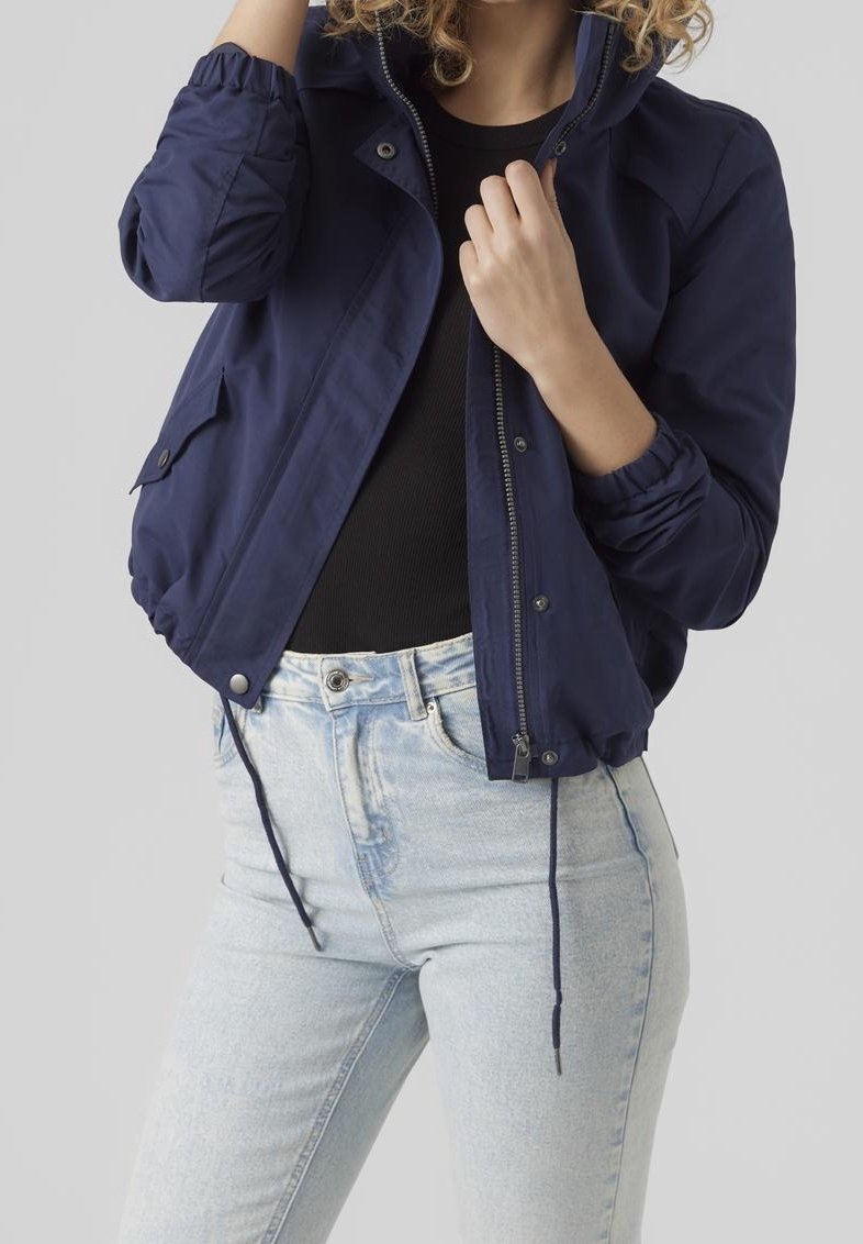 Femme portant une veste zippée bleu marine sur un haut noir avec un jean taille haute bleu clair, ajustant le col de la veste d'une main.
