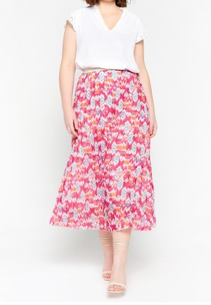 Vrouw met een wit kort mouwen blouse en een kleurrijke, patroonrijke, enkellange rok met nudekleurige sandaalhakken tegen een witte achtergrond.