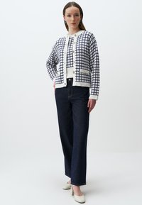 Witte cardigan met marineblauw ruitpatroon, ronde hals en knoopsluiting, gecombineerd met donkerblauwe jeans en witte hoge hakken.