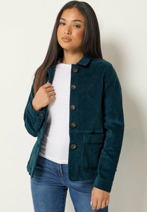 Jeune femme aux longs cheveux foncés porte une veste en velours côtelé turquoise boutonnée par-dessus une chemise blanche et un jean bleu, debout devant un mur clair uni.