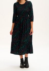 Žena v tmavě zelených midi šatech s pestrými leopardími skvrnami a černými šněrovacími kotníkovými botami stojící na bílém pozadí.