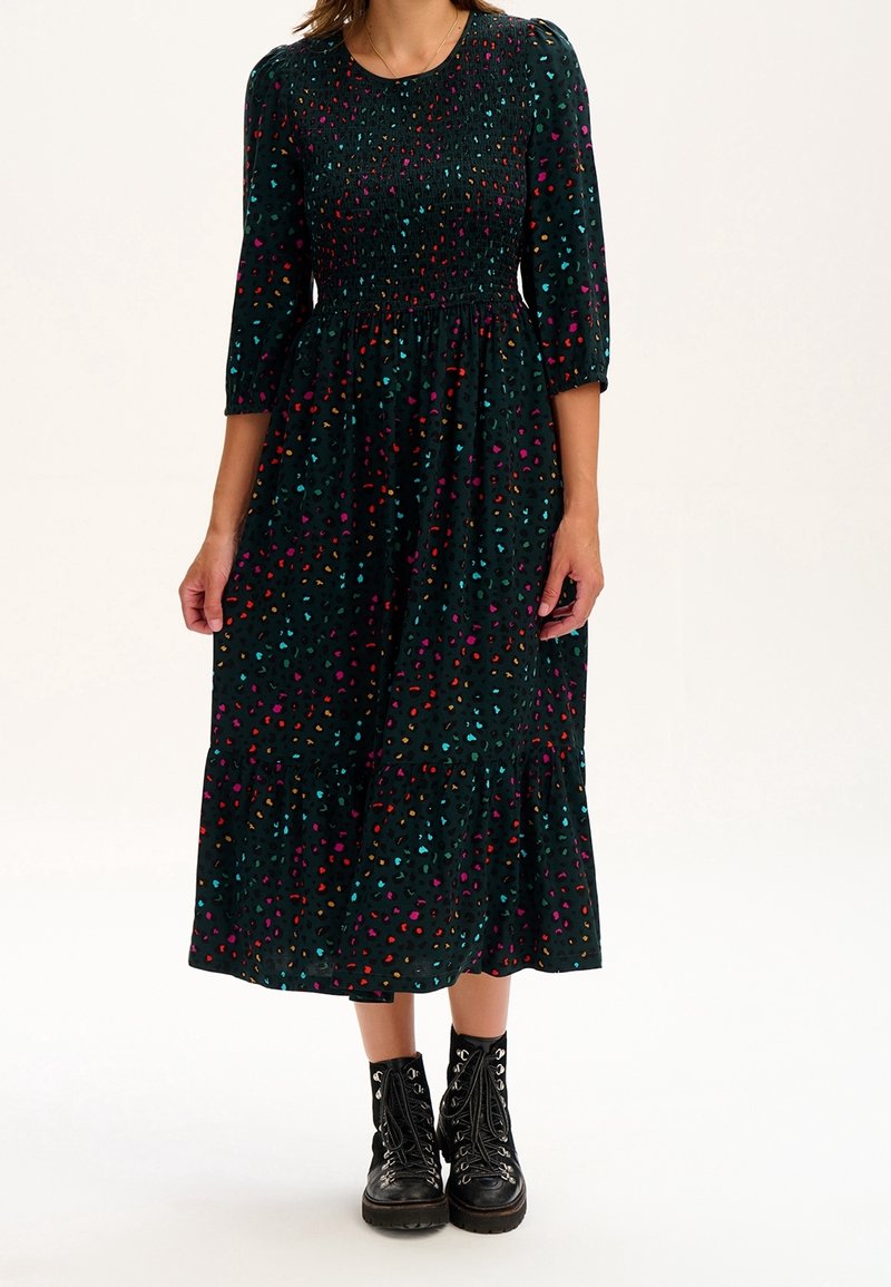 Žena v tmavě zelených midi šatech s pestrými leopardími skvrnami a černými šněrovacími kotníkovými botami stojící na bílém pozadí.