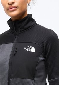 Γυναίκα που φοράει μαύρο και γκρι μπουφάν με φερμουάρ και το λογότυπο The North Face στο στήθος, μπροστά σε λευκό φόντο.