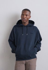 Felpa in cotone blu navy con cappuccio e cordoncino, maniche raglan, tasca frontale e piccolo logo sul petto, abbinata a pantaloni beige.