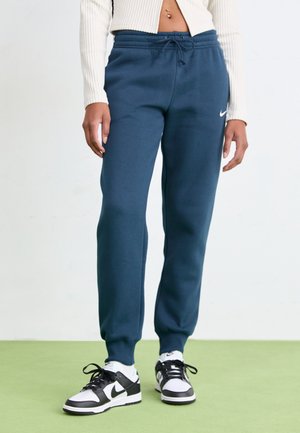 Person trägt blaue Nike-Jogginghose, weißen kurz geschnittenen Pullover und schwarz-weiße Nike-Sneaker und steht auf grünem Boden.