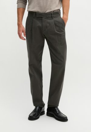 Homme portant un pantalon plissé gris foncé, des chaussures en cuir noires et un haut à manches longues gris clair, debout avec une main dans la poche.