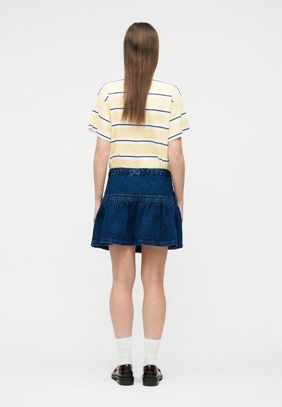 Γυναίκα με μακριά καστανά μαλλιά που φοράει κίτρινη ριγέ μπλούζα, μπλε τζιν φούστα, λευκές κάλτσες και σκούρα μοκασίνια, στέκεται έτσι ώστε να φαίνεται από πίσω, με φόντο λευκό.