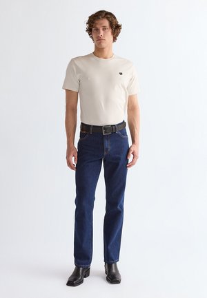 Giovane uomo con capelli ricci che indossa una t-shirt beige a maniche corte, jeans blu scuro con cintura nera e stivaletti neri, in piedi davanti a uno sfondo bianco.