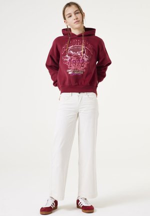Sudadera burdeos con gráfico "Colorado 1972", combinada con pantalones de pierna ancha blancos y zapatillas de gamuza rojas con rayas blancas.