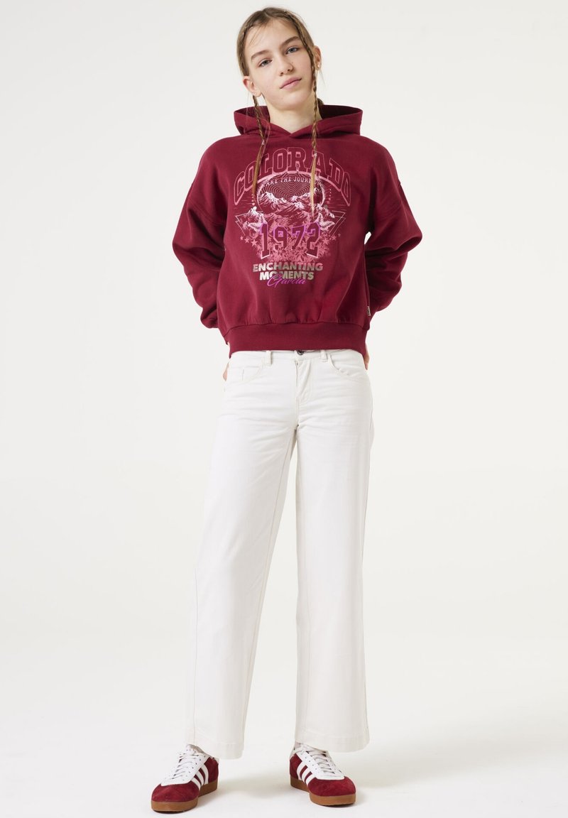 Bordeaux hoodie met "Colorado 1972" graphic, gecombineerd met witte wijde broek en rode suède sneakers met witte strepen.
