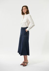 Hvid skjorte med flæsedetaljer, parret med en navy denim midi-nederdel med to forlommer og gule syninger, båret med sorte sandaler.