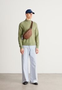 Zöld kötött pulóver, világoskék bőszárú farmer, barna steppelt keresztpántos táska és egy tengerészkék sapka, amelyet egy személy visel egy egyszerű háttér előtt.