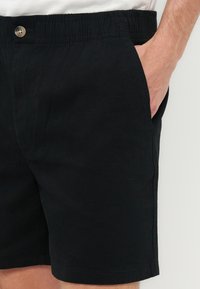 Homme portant un short noir à taille élastique avec un détail de bouton, la main dans la poche droite, et une partie d'une chemise blanche visible.