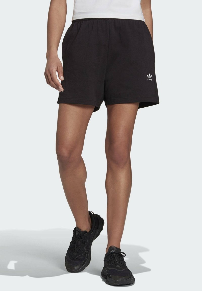 Mustat puuvillahousut, joissa on elastinen vyötärö, ja vasemmassa lahkeessa on pieni valkoinen Adidas-logo, yhdistettynä mustiin urheilukenkäisiin.