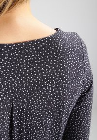 Svart tyg med vita prickar, med rundad halsringning och veckdetalj bak. Slät textur, figursydd design.