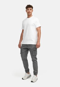 Homme debout portant un T-shirt blanc uni à manches courtes, un pantalon cargo gris avec poches, et des baskets blanches, devant un fond uni.
