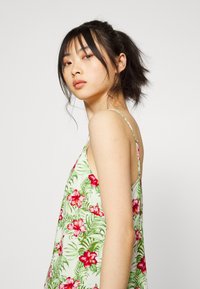Vestido slip floral em verde claro, apresentando hibiscos vermelhos e folhas verdes, com alças finas e um corte descontraído. Textura de tecido suave.