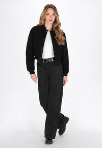 Junge Frau mit langen blonden Haaren, die eine schwarze cropped Jacke, ein weißes Oberteil, weite schwarze Jeans und schwarze Stiefel trägt, während sie nach vorne geht.