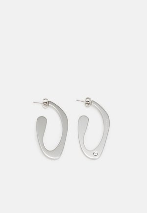 Zilveren metalen oorbellen met een vloeiende, abstracte vorm en een gepolijste afwerking. Één is geaccentueerd met een kleine kristaldetail.