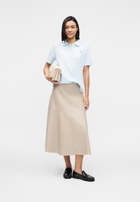 Ανοιχτό μπλε πόλο πουκάμισο με κοντά μανίκια, μεσαίο μπεζ φούστα από συνθετικό δέρμα και μαύρα μοκασίνια, κρατώντας μια μπεζ τσάντα. Καθαρός σχεδιασμός και λεία υφή.