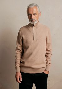 Jasno beżowy sweter o dzianinowej strukturze z wysokim kołnierzem i pół-zapięciem na guziki. Posiada ściągacze na mankietach i dolnym brzegu, wykonany z teksturowanej tkaniny.