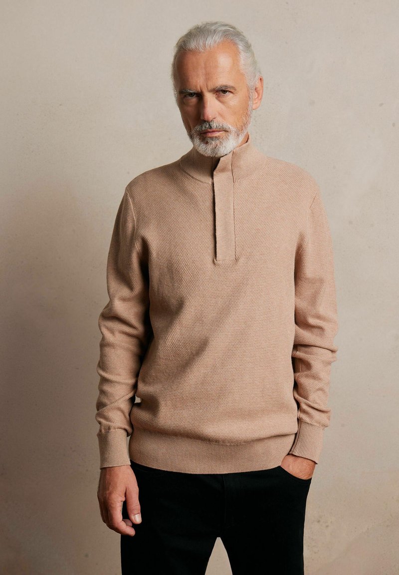 Jasno beżowy sweter o dzianinowej strukturze z wysokim kołnierzem i pół-zapięciem na guziki. Posiada ściągacze na mankietach i dolnym brzegu, wykonany z teksturowanej tkaniny.