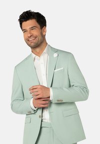 Heller mintgrüner Anzug mit strukturierter Oberfläche, weißem Hemd und einem weißen Blumen-Anstecker. Die Jacke verfügt über zwei Fronttaschen und geknöpfte Manschetten.
