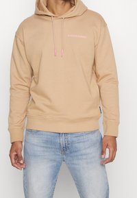 Felpa con cappuccio beige in materiale morbido, con tasca a marsupio frontale, coulisse con dettagli rosa e un piccolo logo sul petto.