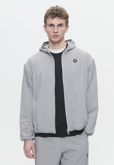 Veste à capuche gris clair avec fermeture éclair sur le devant. Présente un logo noir sur la poitrine gauche et des poches latérales, fabriquée à partir d'un matériau lisse et léger.