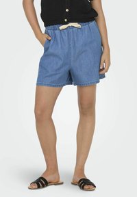 Denim kratke hlače z elastičnim pasom in lahkim materialom, z sprednjimi žepi in kontrastnim detajlom v obliki zaveze, nosene s črnimi natikači.