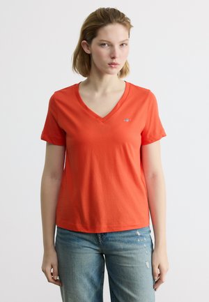 Νεαρή γυναίκα που φοράει απλό πορτοκαλί μπλουζάκι με V λαιμόκοψη και κοντά μανίκια, καθώς και ανοιχτόχρωμο μπλε τζιν παντελόνι, μπροστά σε ουδέτερο φόντο.
